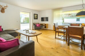 Een woonkamer met een bank, een salontafel, een eettafel met stoelen, een televisie en een raam met uitzicht op groen.