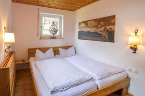 A double bed with white linens in a room with wooden beams on the ceiling, a window with a leaf-shaped decoration, and a painting of a landscape with cows. - Holiday apartment with 3 bedrooms (Garmisch-Partenkirchen)