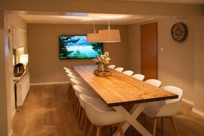 A dining area with a long wooden table, white chairs, a television mounted on the wall, and a clock on the wall.