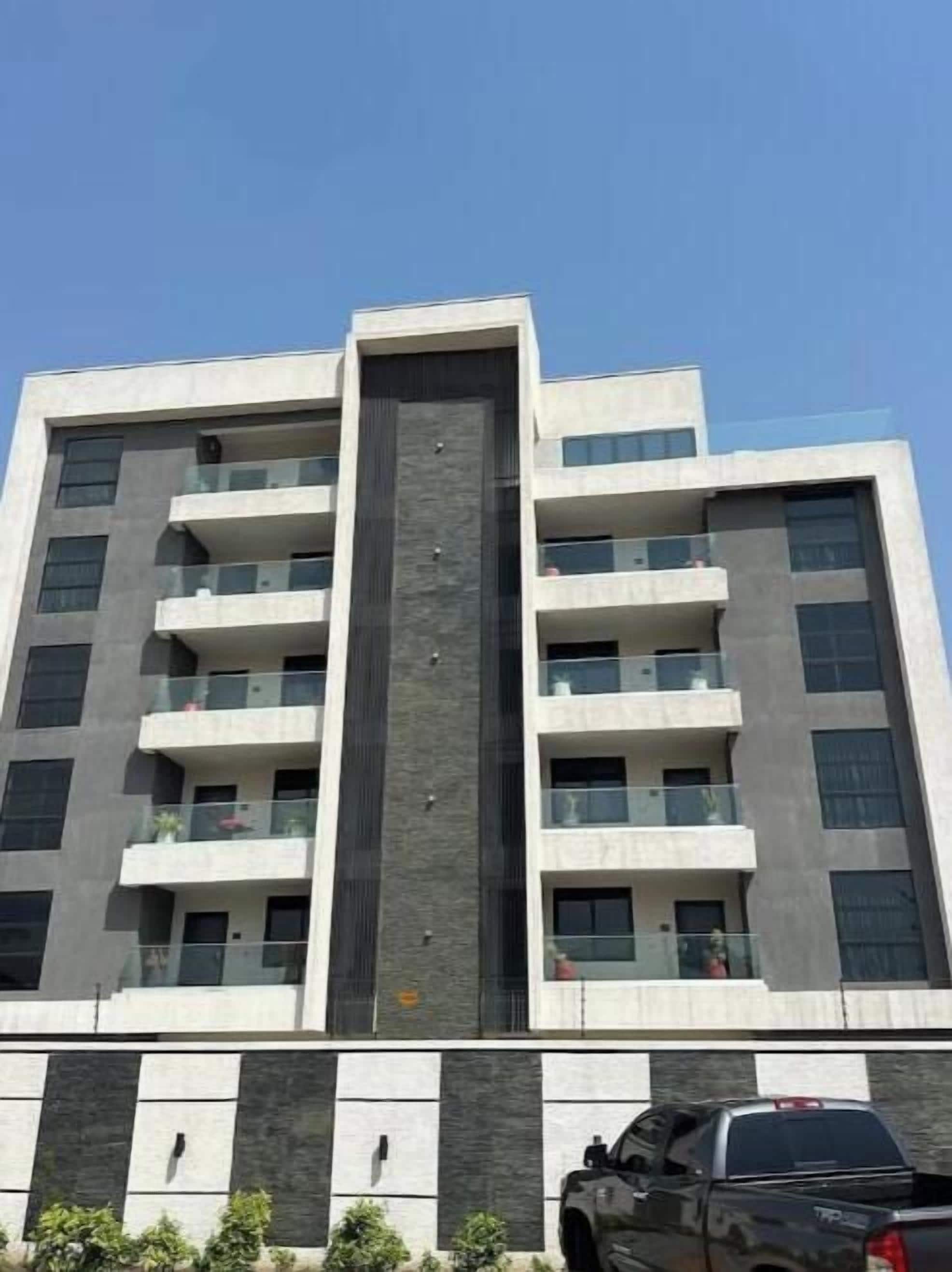 A multi-story building with balconies, a central vertical feature, and a parking area with a truck.