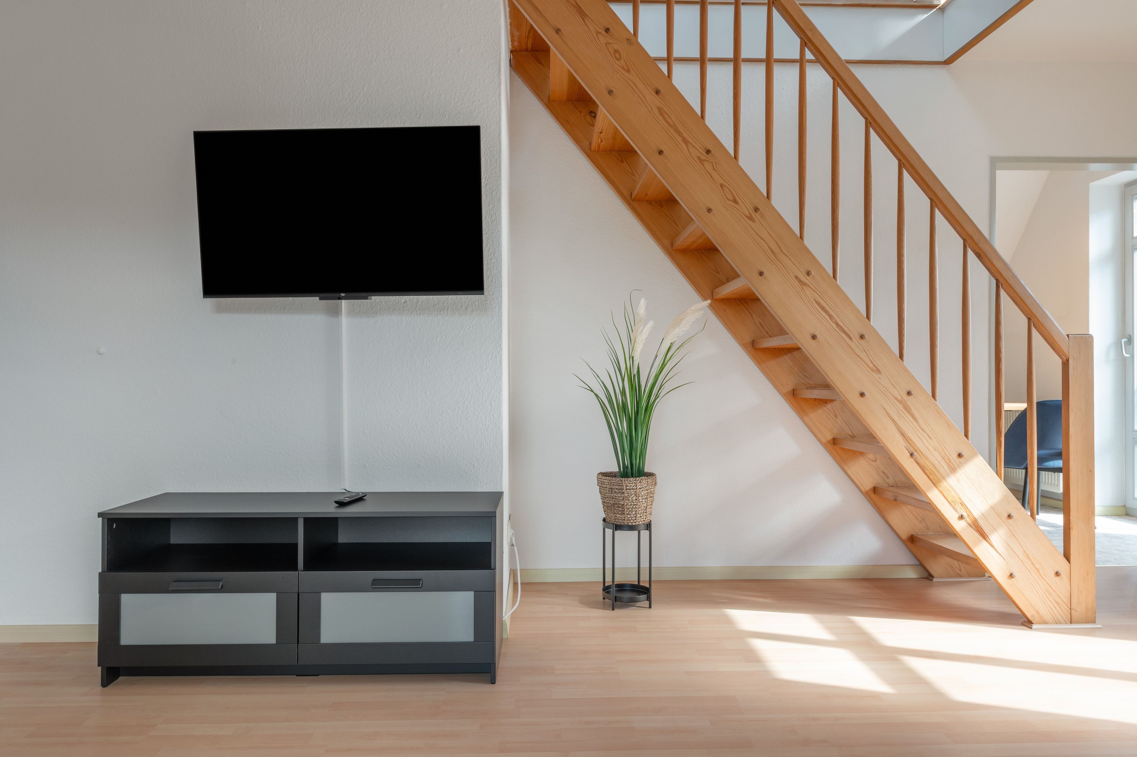 A modern living room with a flat-screen TV mounted on the wall, a wooden staircase, and a small potted plant on a stand.