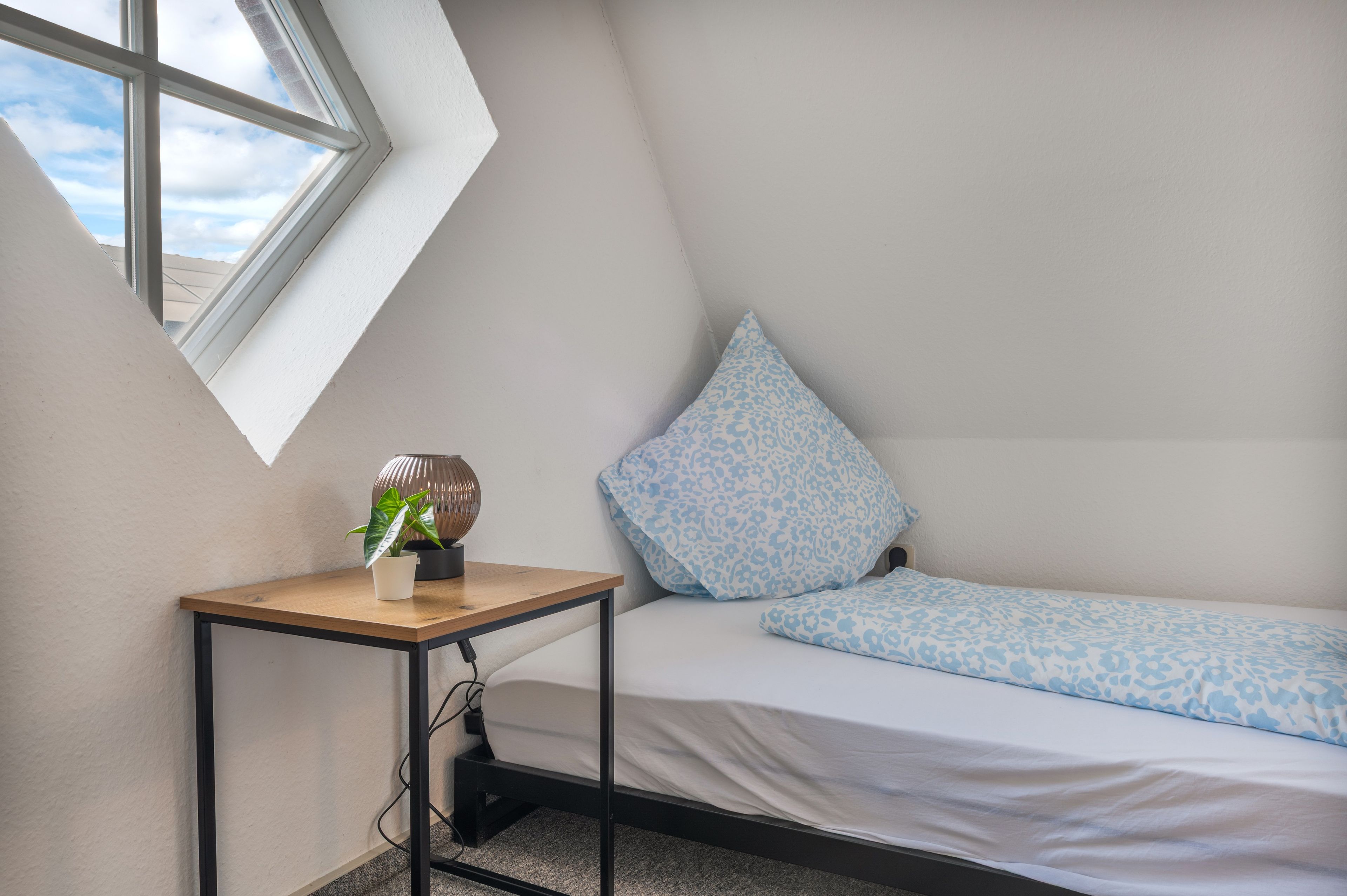 A small, triangular room with a bed, a table with a plant, and a window.