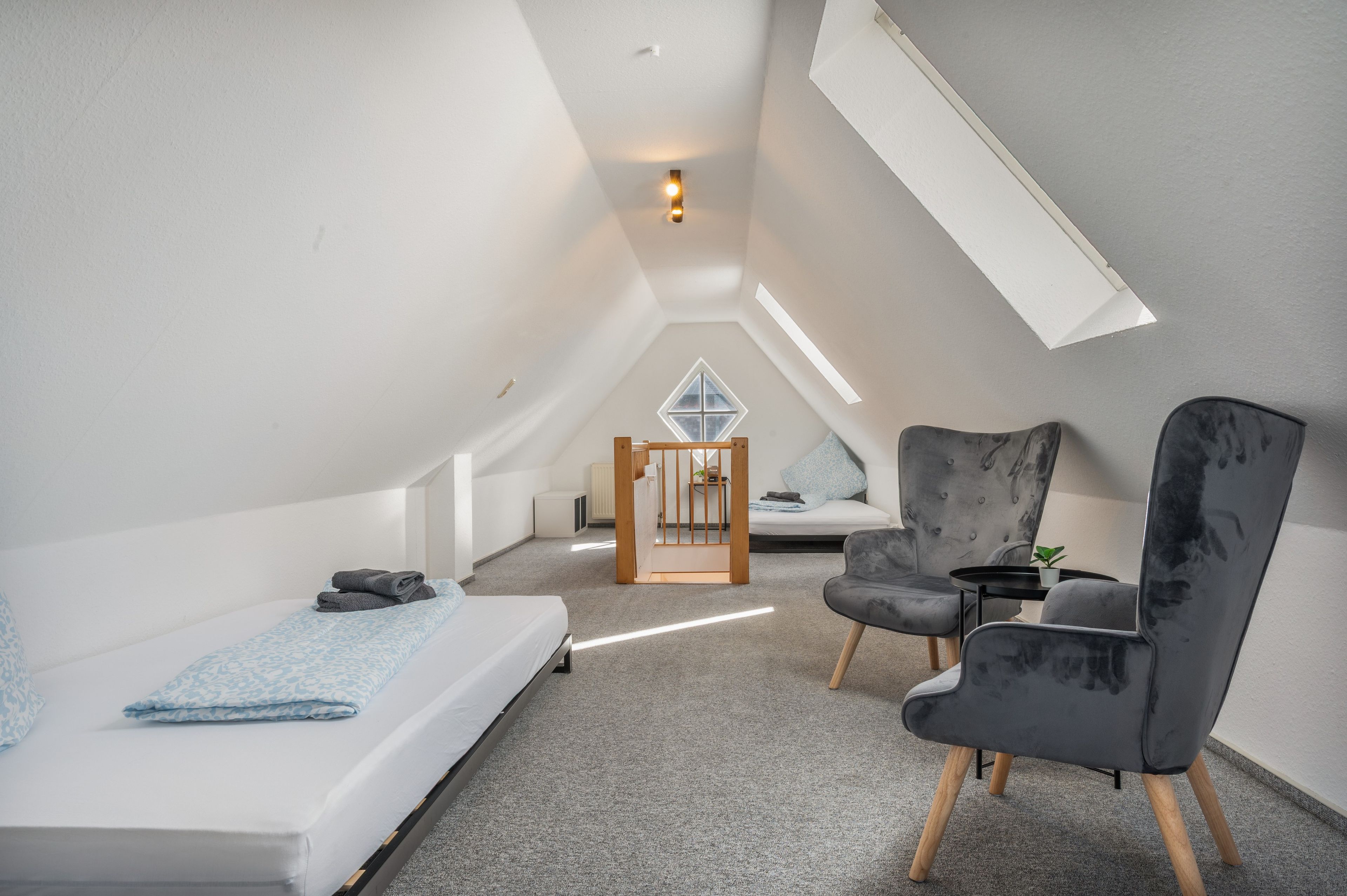 A modern loft bedroom with a sloped ceiling, a skylight, a bed with a patterned bedspread, a seating area with a chair and a small table, and a view of the adjacent room through a window.