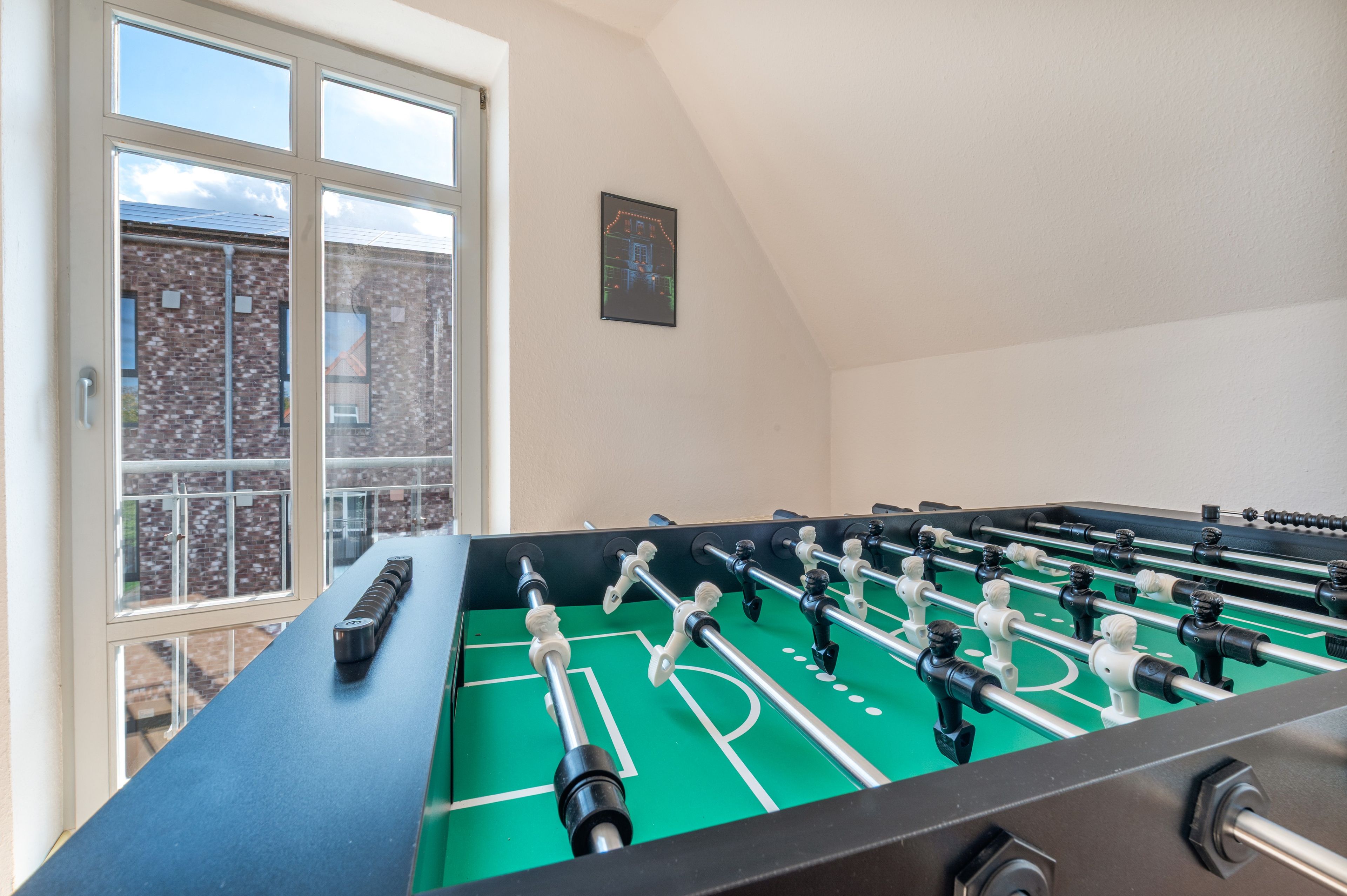 Een tafelvoetbaltafel in een ruimte met een groot raam, een lijst met een foto aan de muur, en een bakstenen gebouw buiten.