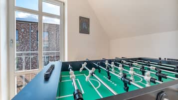 Een tafelvoetbaltafel in een ruimte met een groot raam, een lijst met een foto aan de muur, en een bakstenen gebouw buiten.