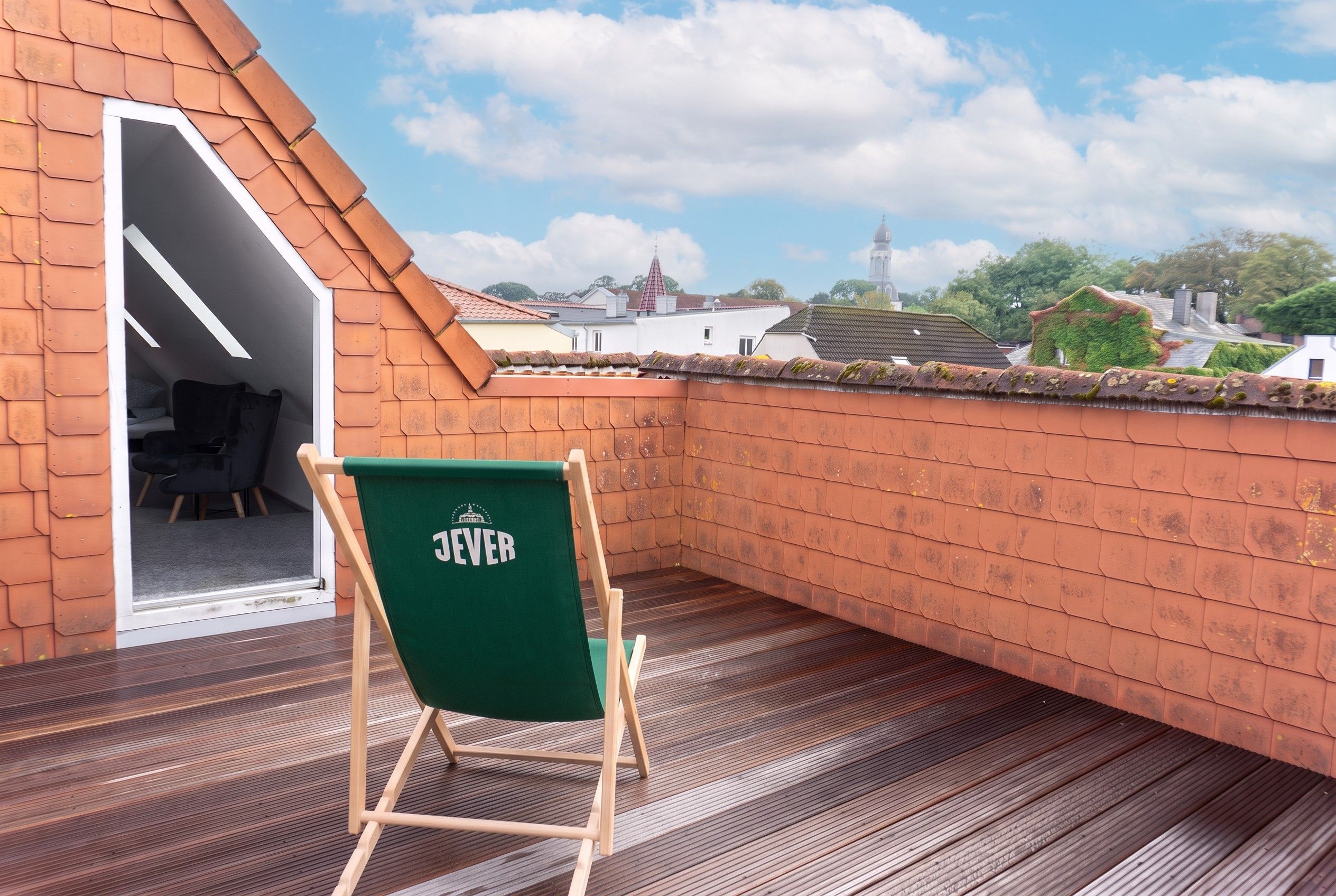 En takterrass med en grön stol märkt med texten ”JEVER”, trägolv och utsikt över byggnader och ett torn i fjärran.