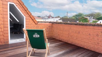 Una terraza en la azotea con una silla verde identificada con 'JEVER', suelo de madera y vistas a edificios y una torre a lo lejos.