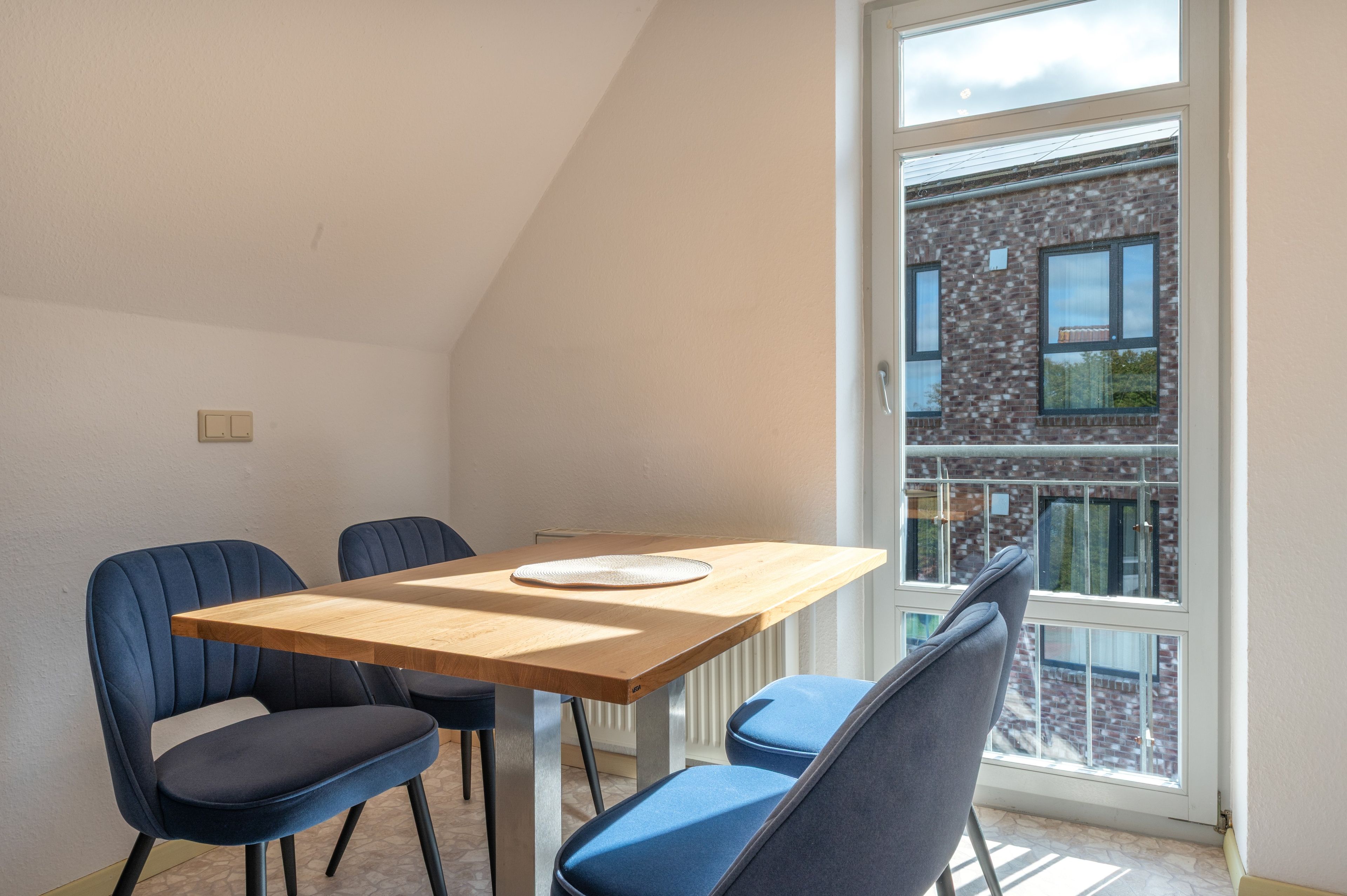 A dining area with a wooden table and four chairs, a window with a view of a brick building, and a white ceiling.