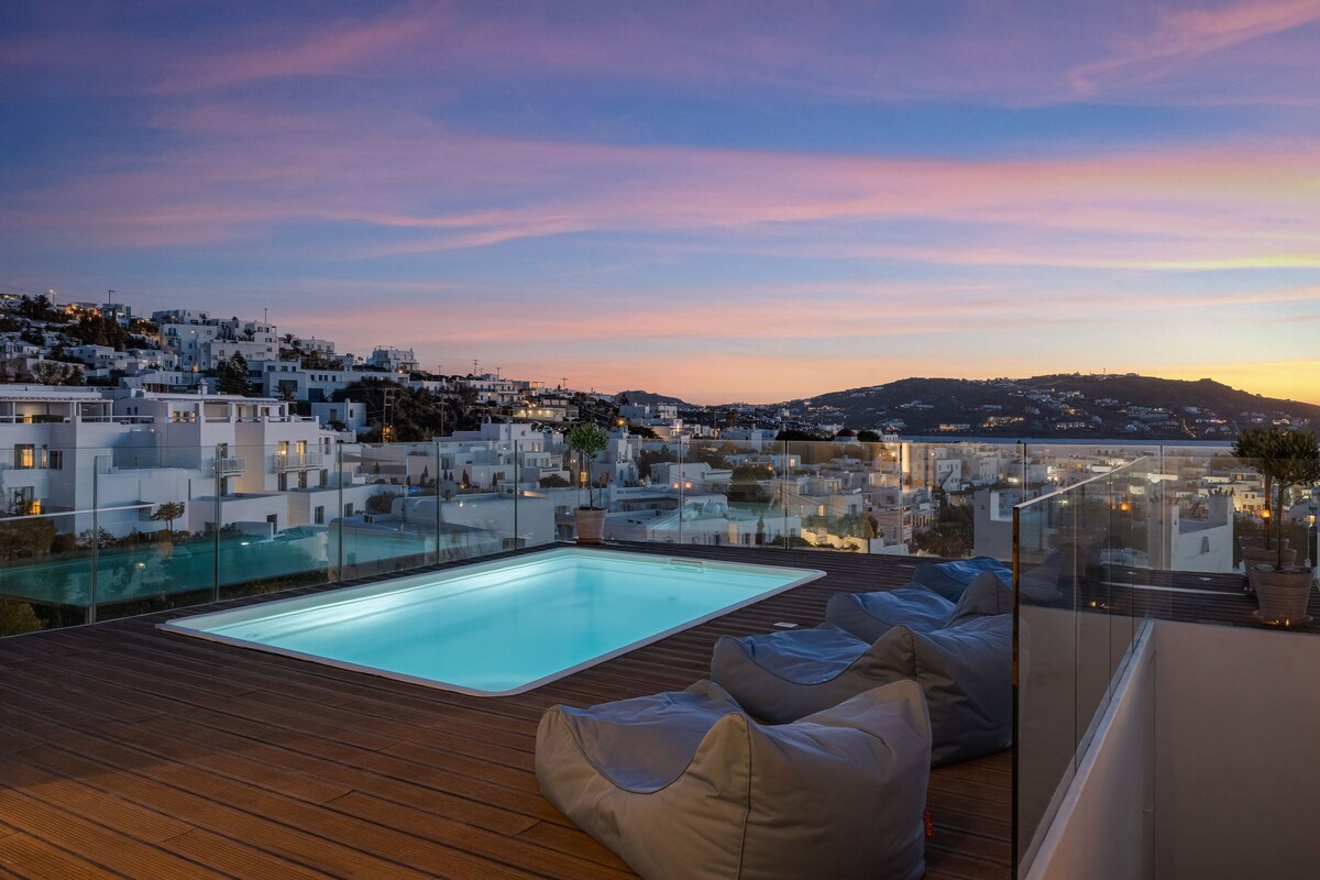 A rooftop pool area with a glass barrier, a wooden deck, and a view of a coastal town at dusk.