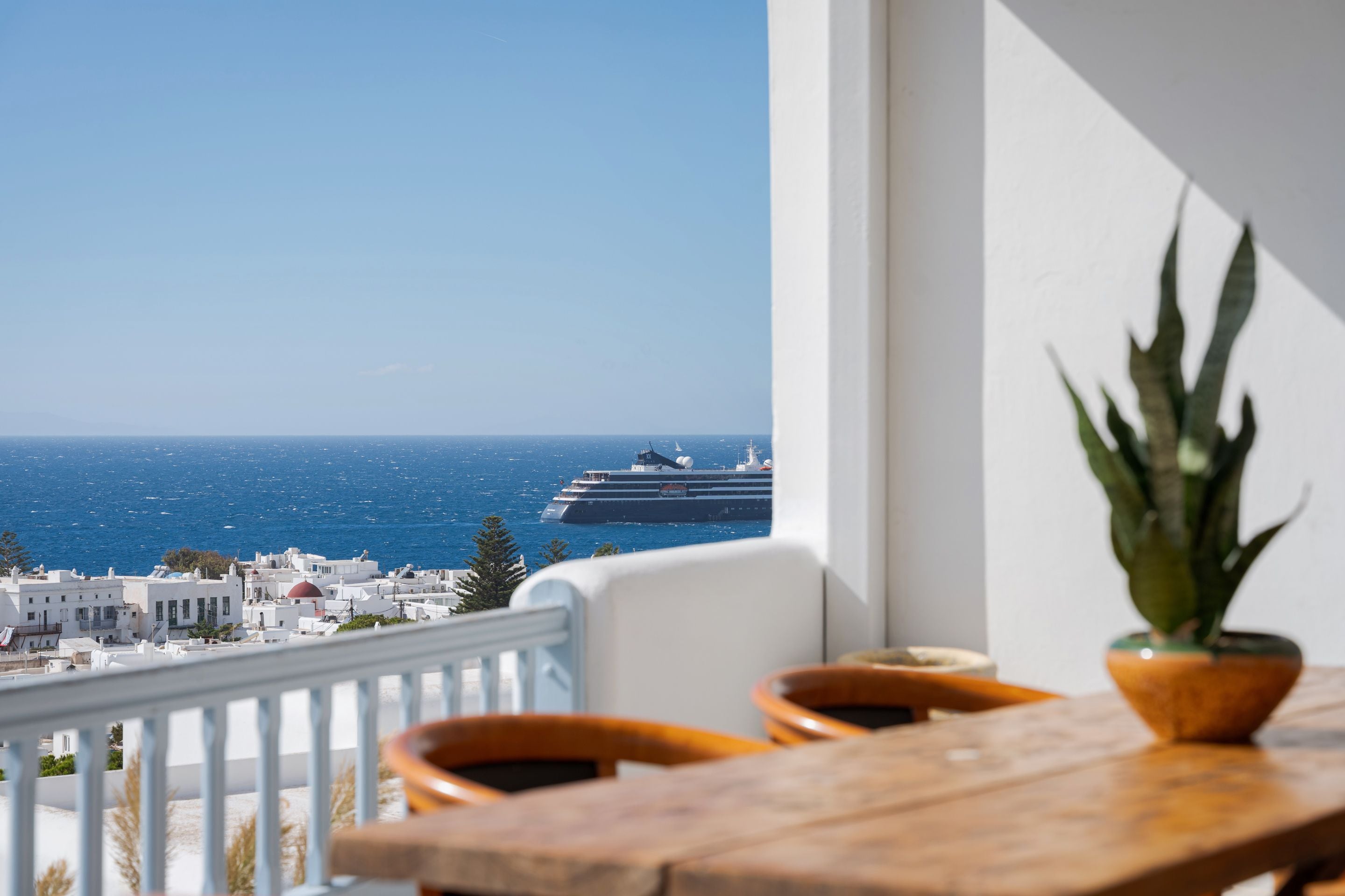 Ein Balkon mit Blick auf das Meer und ein Kreuzfahrtschiff, weiße Gebäude und ein Blumentopf.