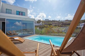 A modern outdoor swimming pool with wooden loungers, a glass-enclosed area, and a view of a hillside town.