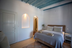 A bedroom with a bed, bedside tables, a nightstand, a door, and an arched doorway. - Vastao Luxury Residences (Mykonos)