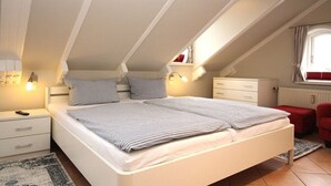 Een slaapkamer met een schuine plafond, een bed met gestreept beddengoed, bedlampen, een nachtkastje, een dressoir en een raam met gordijnen.