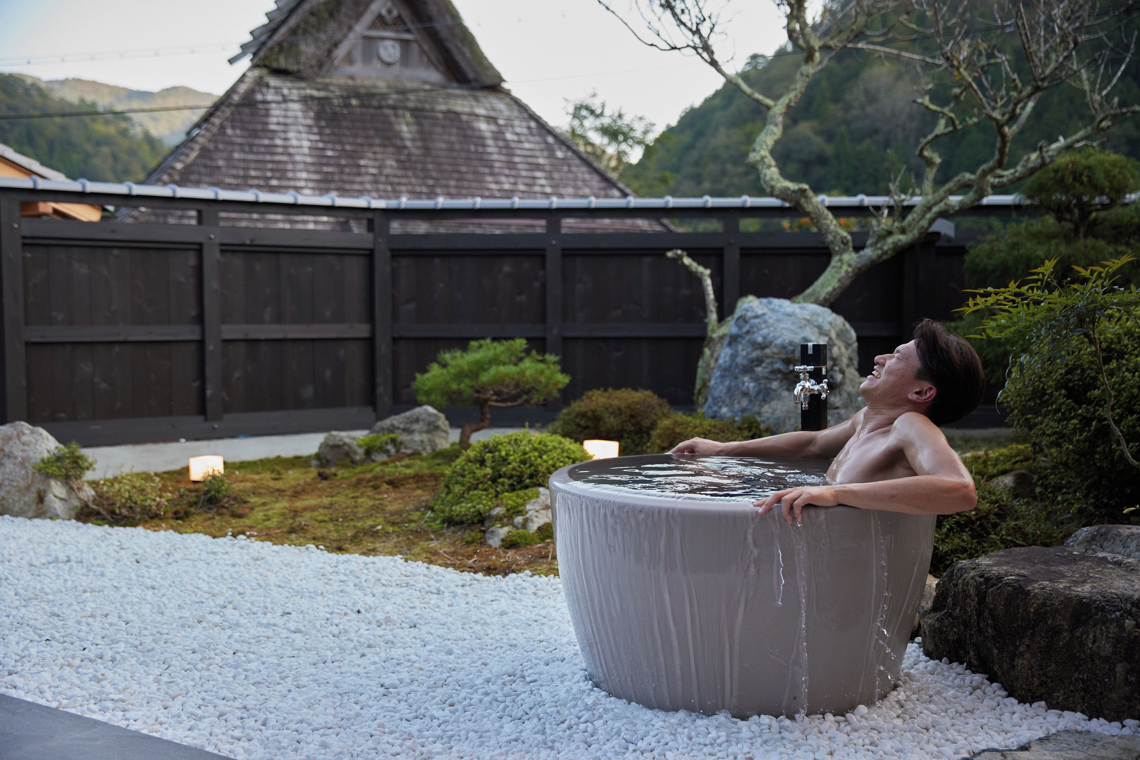 A person relaxing in a hot tub surrounded by white pebbles, with a traditional building and a wooden fence in the background.