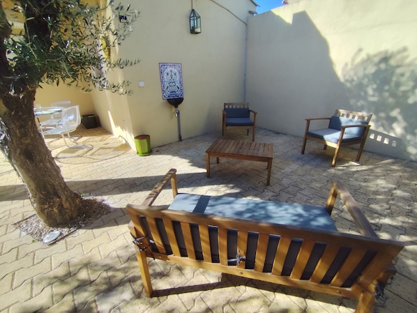 Un patio con un banco de madera, dos sillas, una mesita y un árbol.