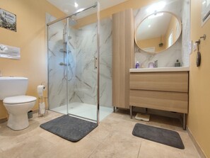 Un baño con ducha envuelta en vidrio, inodoro blanco, tocador de madera con espejo y salpicadero de mármol.