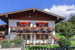A chalet with a balcony adorned with flowers and two red umbrellas, surrounded by a fenced garden and a mountain backdrop. - Holiday apartment with 1 bedroom (Schönau am Königssee)
