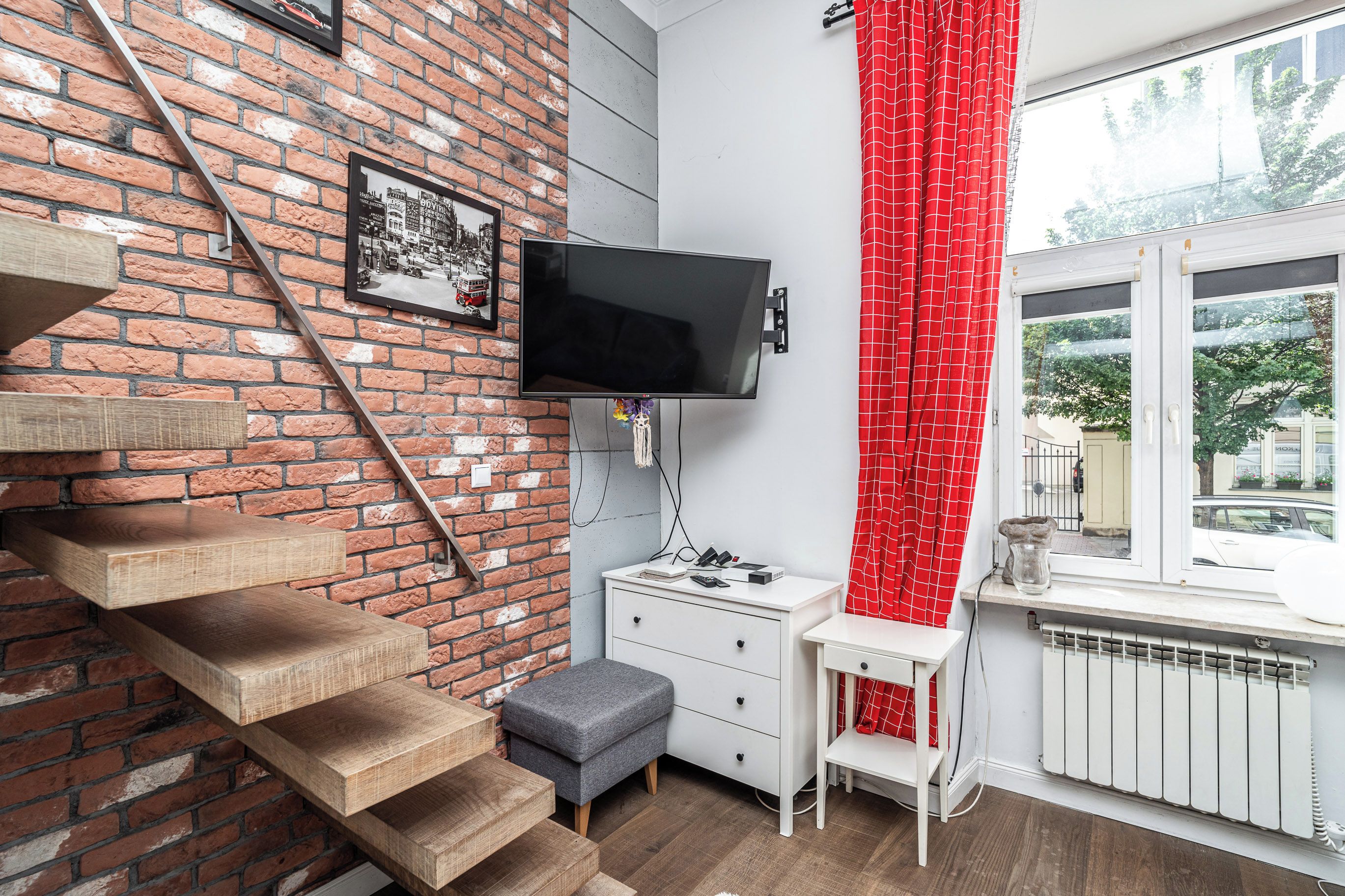 A room with a brick wall, a flat-screen TV mounted on the wall, a white desk with drawers, a grey ottoman, and a window with a red curtain.