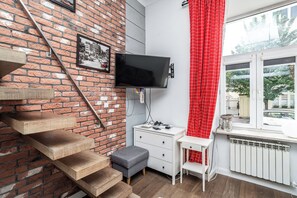 Et værelse med murstensvæg, et fladskærms-tv ophængt på væggen, et hvidt skrivebord med skuffer, en grå skammel og et vindue med rødt gardin.