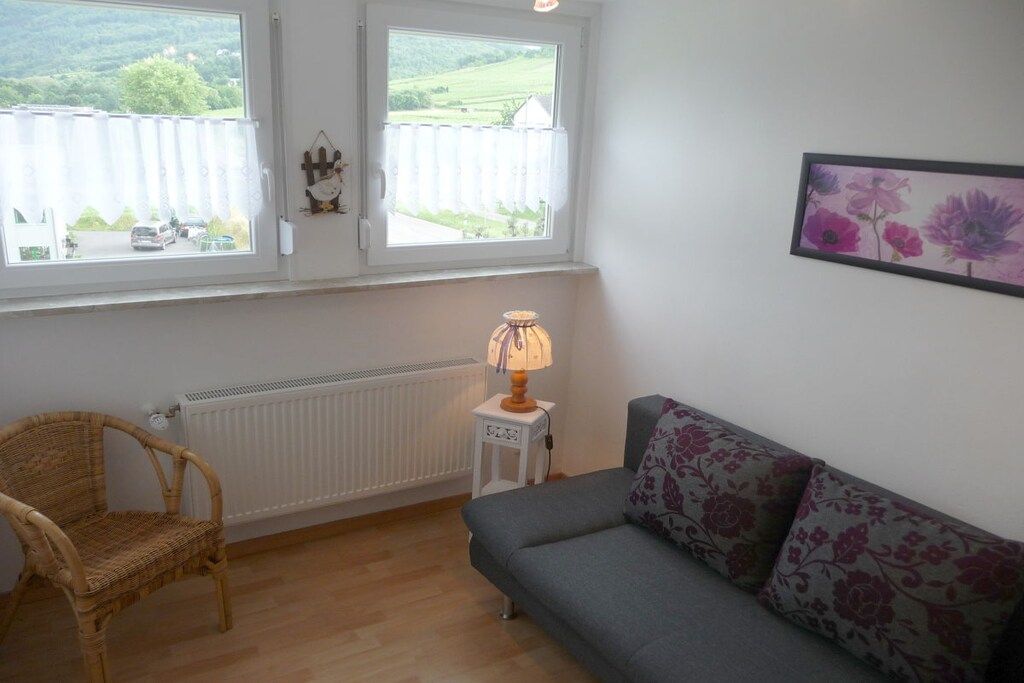 Et værelse med en grå sofa, en fletstol, et lille bord med en lampe, en radiator og et vindue med udsigt til grøn bevoksning.