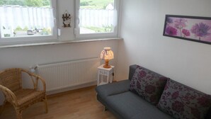 A room with a grey sofa, a wicker chair, a small table with a lamp, a radiator, and a window with a view of greenery. - Holiday apartment with 2 bedrooms (Traben-Trarbach\/Wolf)