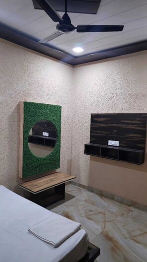A room with a bed, a ceiling fan, a green wall with a circular mirror, and a shelf with a black surface. - Dream In (Mathura)