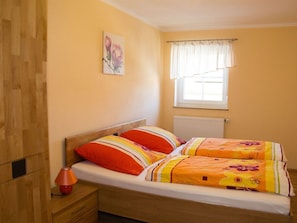 A single bed with a wooden headboard, a bedside table with a lamp, and a window with a white curtain. - 3 - Schuepferlingshof (Kunreuth)