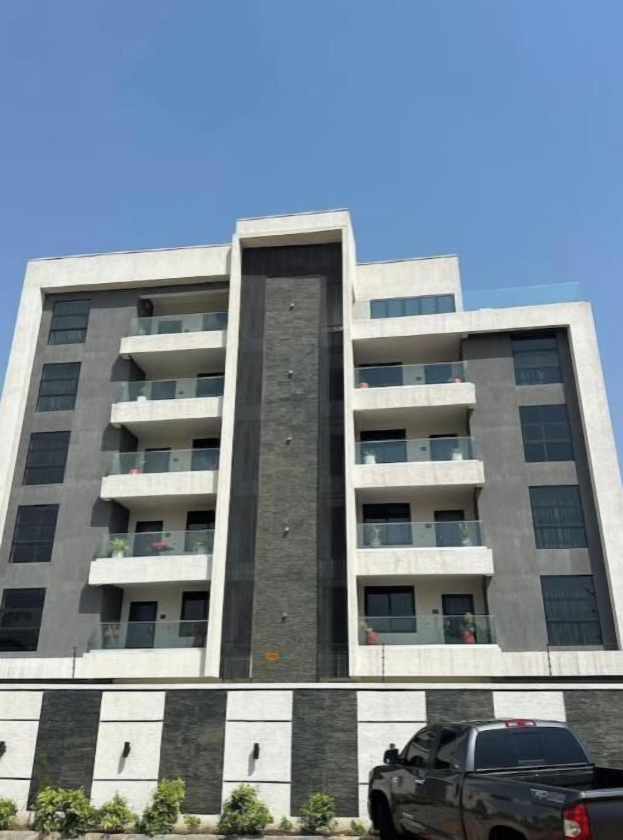 A multi-story building with balconies, a central vertical feature, and a parking area with a truck.