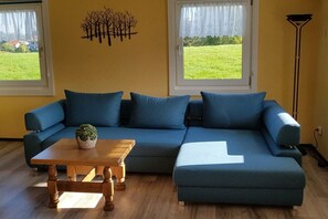 A living room with a blue sofa, a wooden coffee table, and a potted plant. There are two windows with curtains, and a floor lamp is visible. - Holiday apartment with 3 bedrooms (Röthenbach (Allgäu))