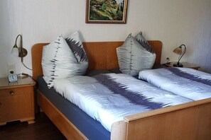 A double bed with a blue mattress and two pillows with a leaf pattern. There is a wooden headboard, a bedside table with a lamp, and a wall clock. - Holiday apartment with 3 bedrooms (Röthenbach (Allgäu))