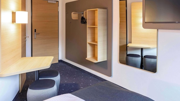 A modern hotel room with a wooden desk, a small seating area, a built-in shelf, and a flat-screen TV mounted on the wall. - B&B HOTEL Fürth-Hardhöhe (Fürth)