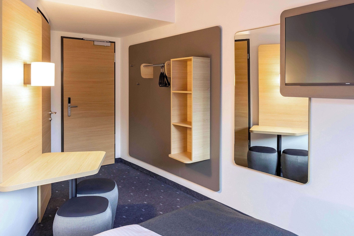 Moderni hotellihuone, jossa on puinen työpöytä, pieni oleskelualue, seinään integroitu hylly ja seinälle asennettu litteänäyttöinen televisio.