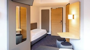 A compact hotel room with a single bed, a bedside table, a small stool, a mirror, and a TV mounted on the wall. - B&B HOTEL Fürth-Hardhöhe (Fürth)