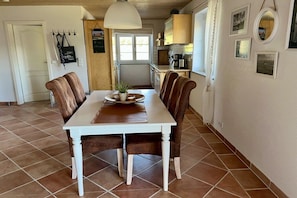 A dining area with a white table and brown chairs, a hanging light fixture, and a kitchen in the background. - 1 in the Walser country house (Wasserburg)