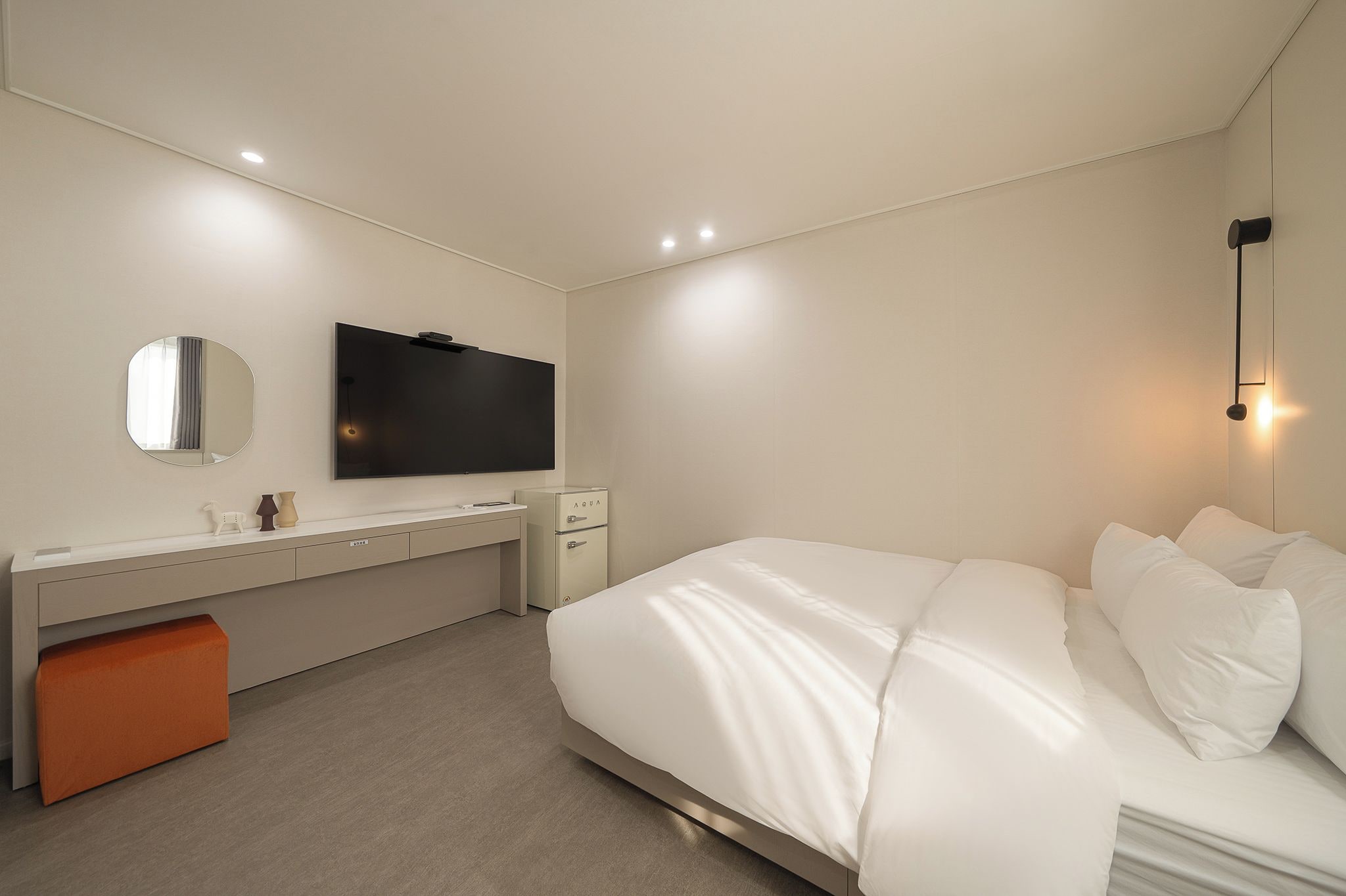 Una habitación de hotel moderna con una cama grande, un escritorio con televisor de pantalla plana, un pequeño taburete naranja y una mesita de noche con lámpara.