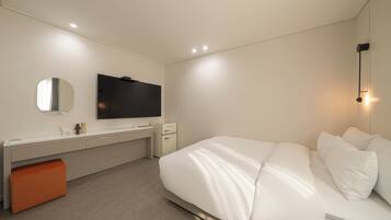 Una habitación de hotel moderna con una cama grande, un escritorio con televisor de pantalla plana, un pequeño taburete naranja y una mesita de noche con lámpara.