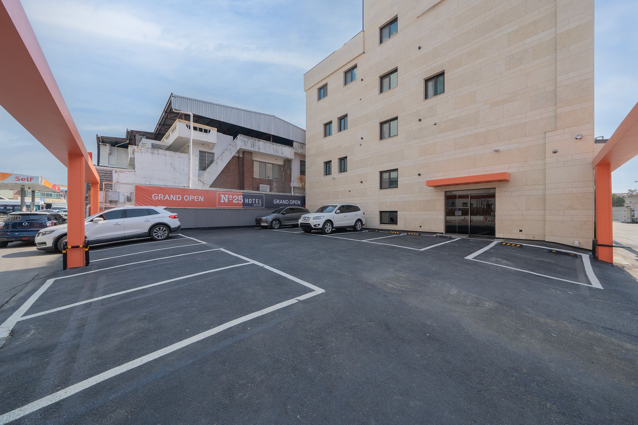 A hotel parking lot with marked spaces, a building with a sign indicating a grand opening, and a self-service fuel station in the background.