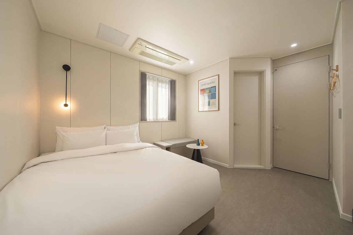 A hotel room with a large bed, a bedside table, a small round table, a window with curtains, a door, and a wall-mounted air conditioner.