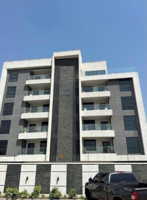 Um edifício com vários andares, varandas, um elemento vertical central e uma área de estacionamento com um veículo.