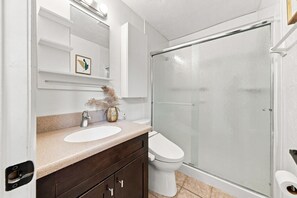 Un bany amb un lavabo blanc, un tocador de fusta fosca, un vàter i una dutxera amb envà.