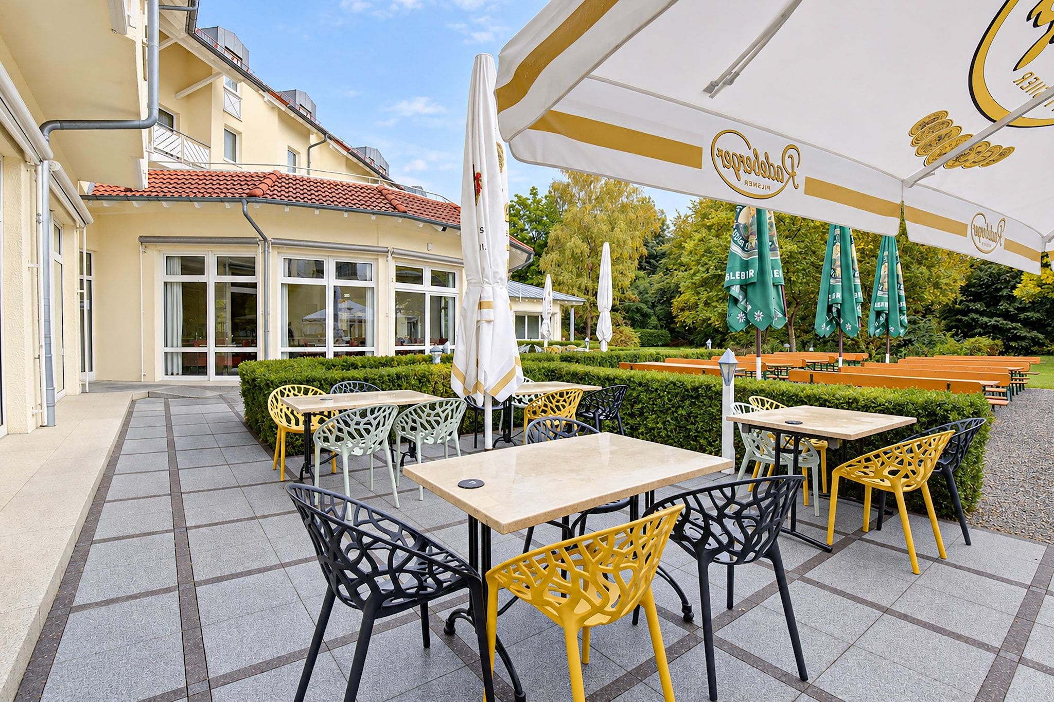 Ein Sitzbereich im Freien mit Tischen und Stühlen, ein überdachter Bereich mit Sonnenschirmen und ein Gebäude mit großen Fenstern im Hintergrund.