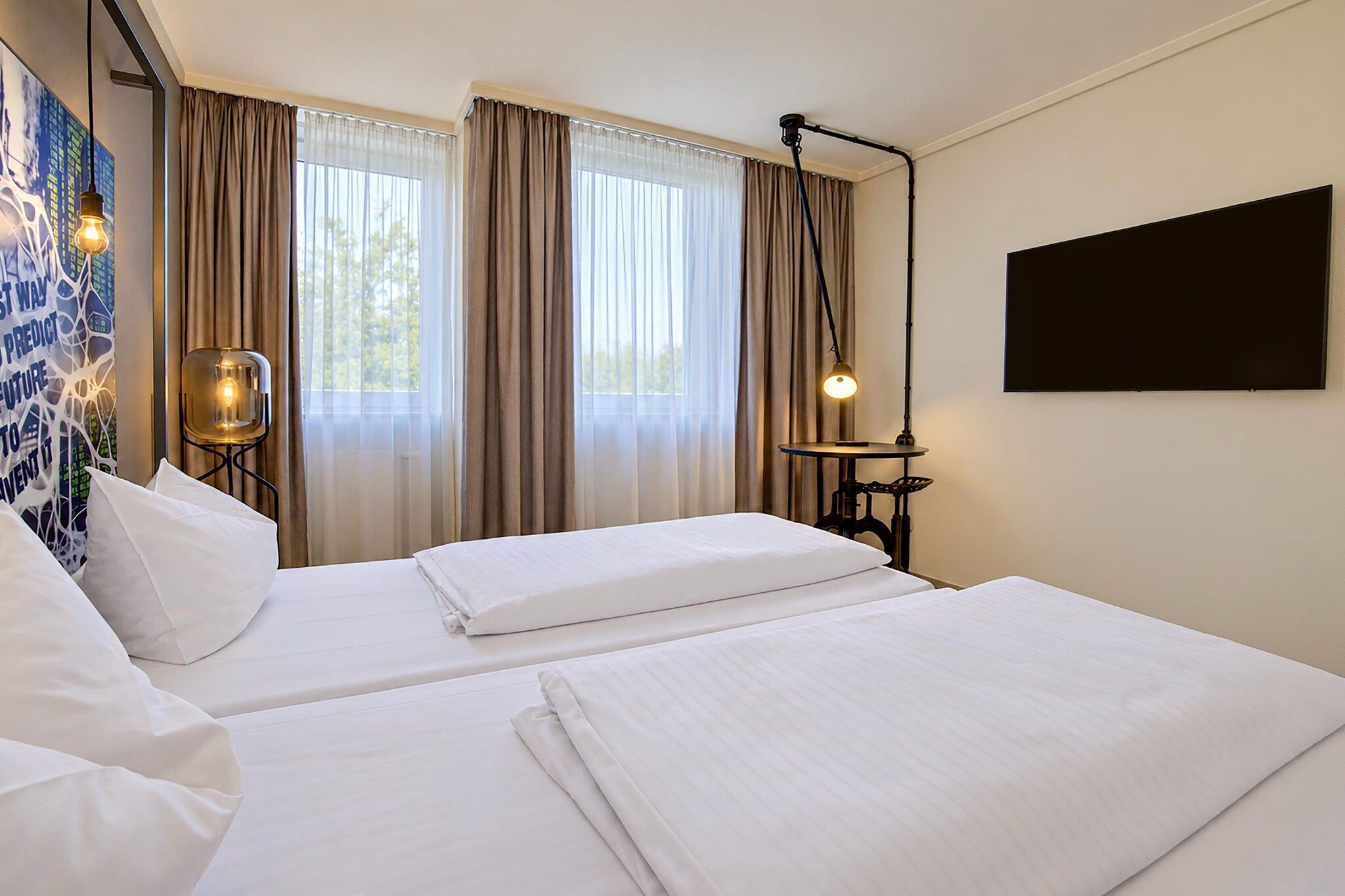 Ein Hotelzimmer mit zwei Betten, einem großen Fenster mit Vorhängen, einem an der Wand befestigten Fernseher und einem Tisch mit einer Lampe.