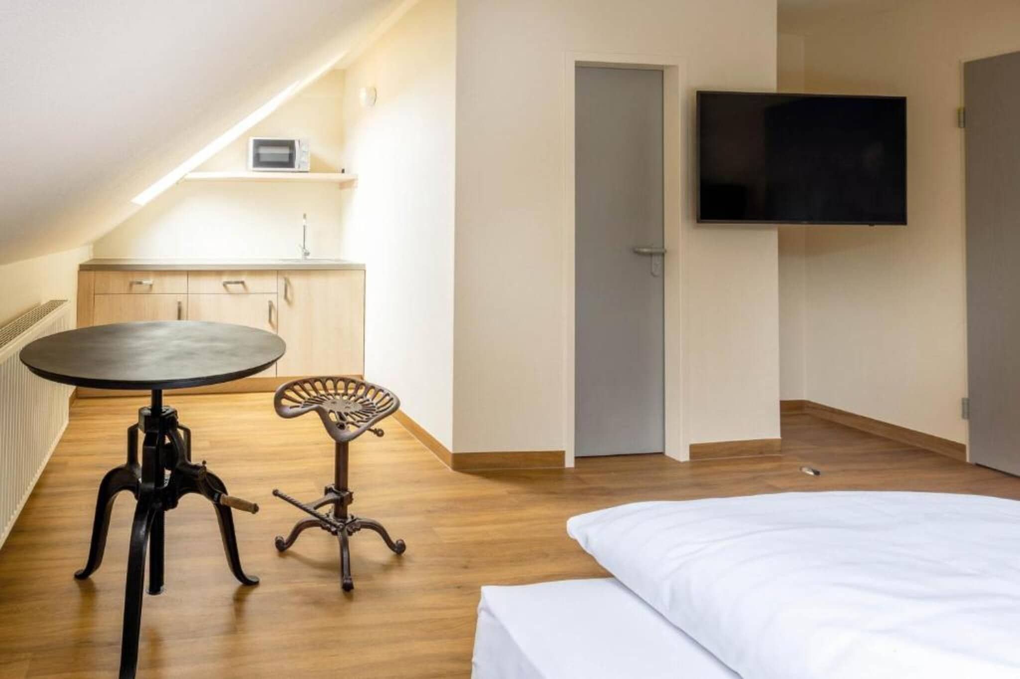 Un dormitorio con cama, una mesita, una silla, televisor y una kitchenette al fondo.