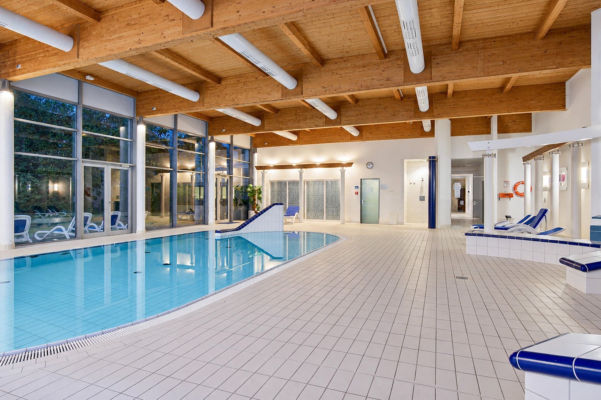 Ein Innenpool mit großen Glaswänden, Fliesenboden und Holzbalkendecke.