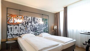 Una habitación de hotel con cama, una mesita, una obra de arte en la pared y una ventana con cortinas.