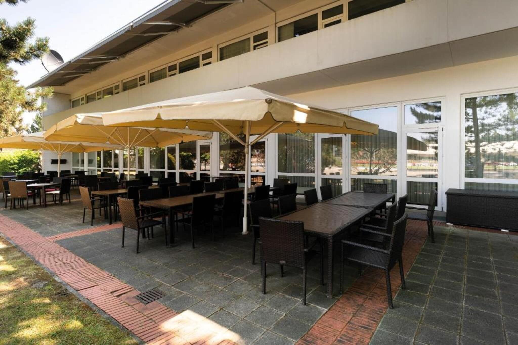 Un área de patio al aire libre con mesas y sillas, cubierta por sombrillas grandes, y un edificio con amplias ventanas al fondo.