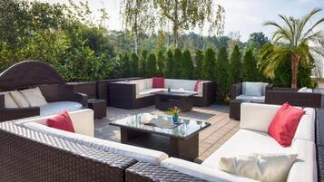 An outdoor seating area with wicker furniture, a glass coffee table, and a view of a forested hillside.