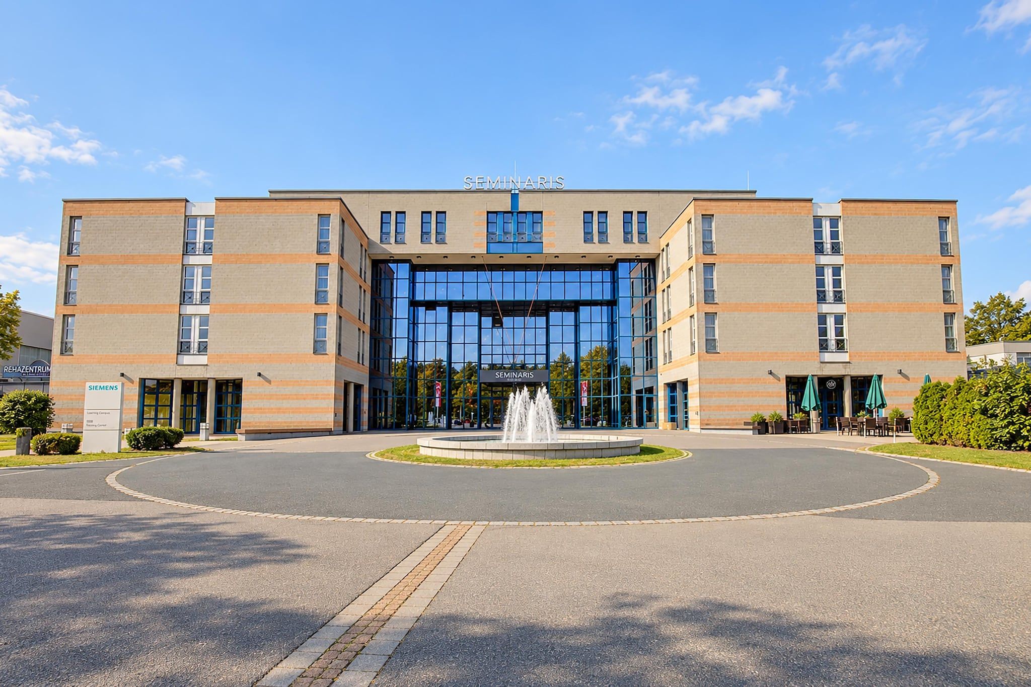 Ein modernes Gebäude mit einem zentralen Eingang, einem Brunnen und blauem Himmel.
