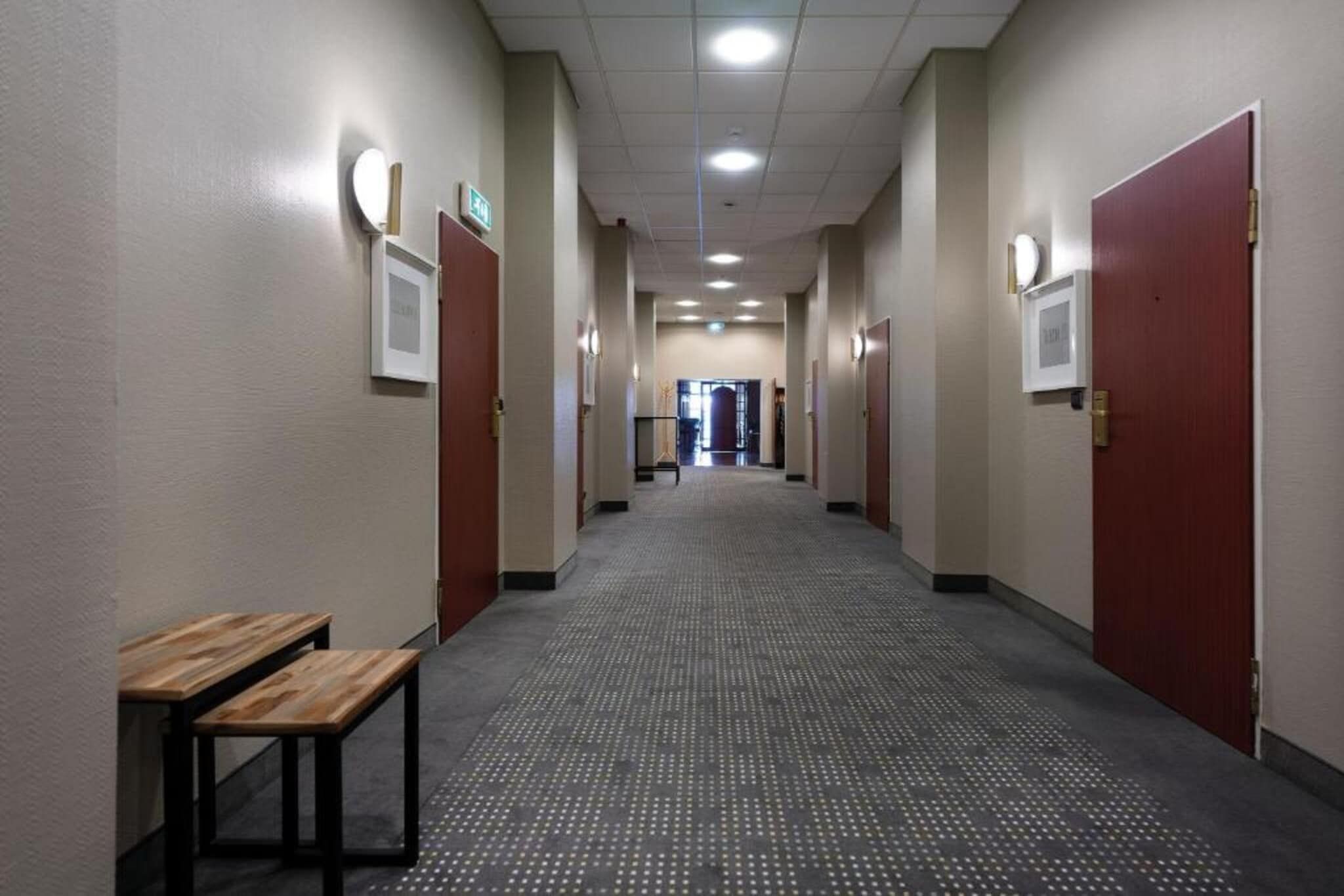 A hotel corridor with a wooden bench, doors, and a carpeted floor.