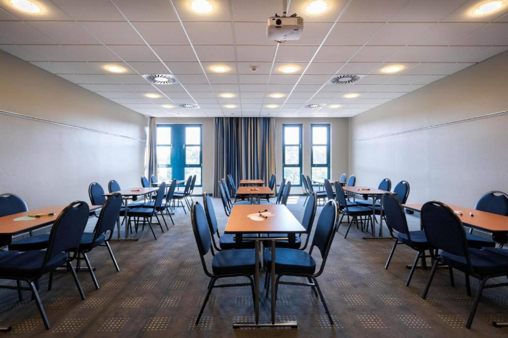 Ein Konferenzraum mit mehreren Tischen und Stühlen, großen Fenstern und Deckenprojektoren.